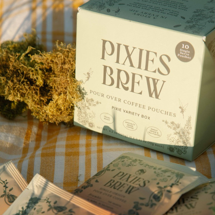 Close-up of a Pixies Brew 'Pixie Variety Box' resting on a yellow gingham picnic blanket in warm sunlight, accompanied by dried moss and scattered sachets
