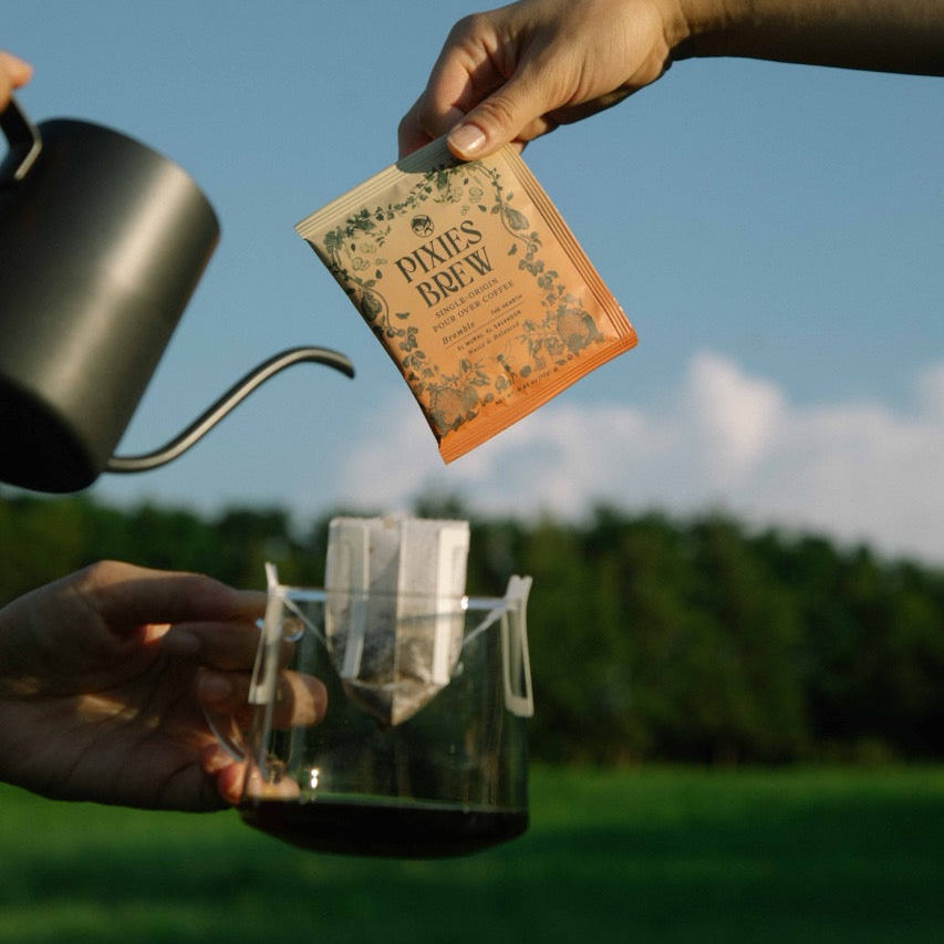 Outdoor coffee preparation scene featuring hands holding a Pixies Brew 'Bramble' sachet and a glass server with a drip filter against a sunny blue sky