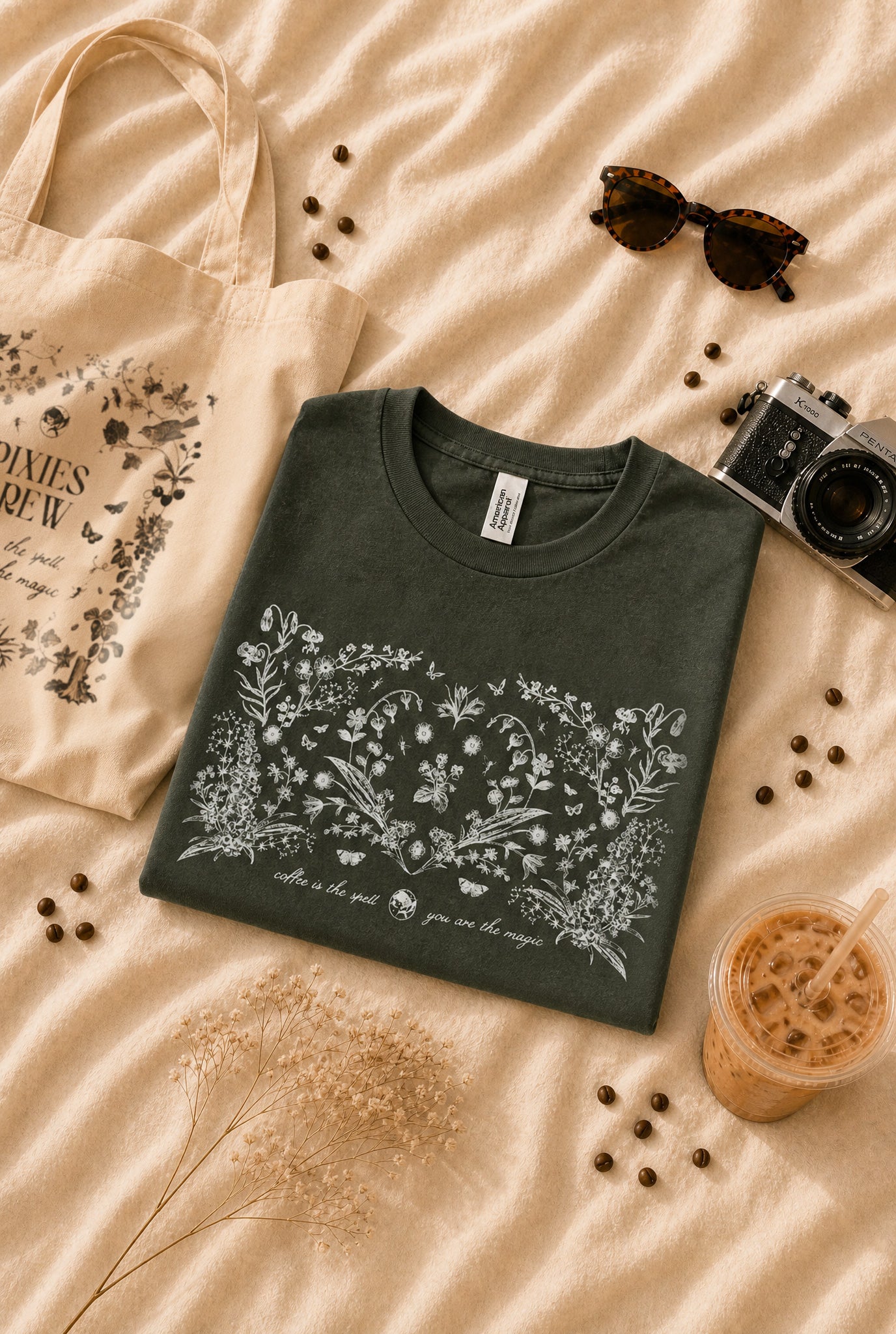 A folded dark green t-shirt with a white floral graphic and text print, styled in a flat-lay arrangement on beige fabric alongside a tote bag, sunglasses, a camera, coffee beans, and an iced coffee drink.