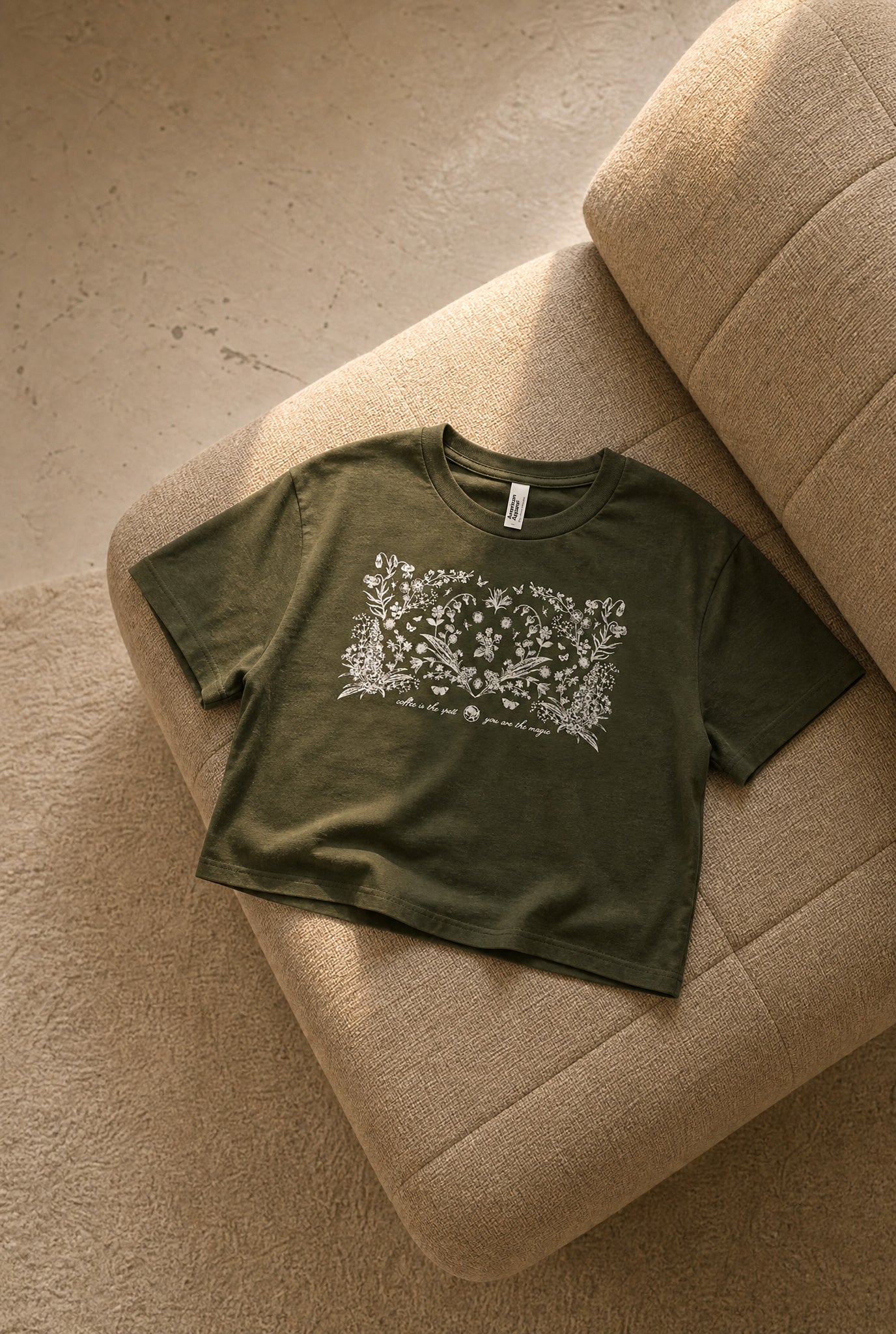 An olive green short-sleeve crop top with a white floral graphic print across the chest, laid flat on a beige textured armchair.