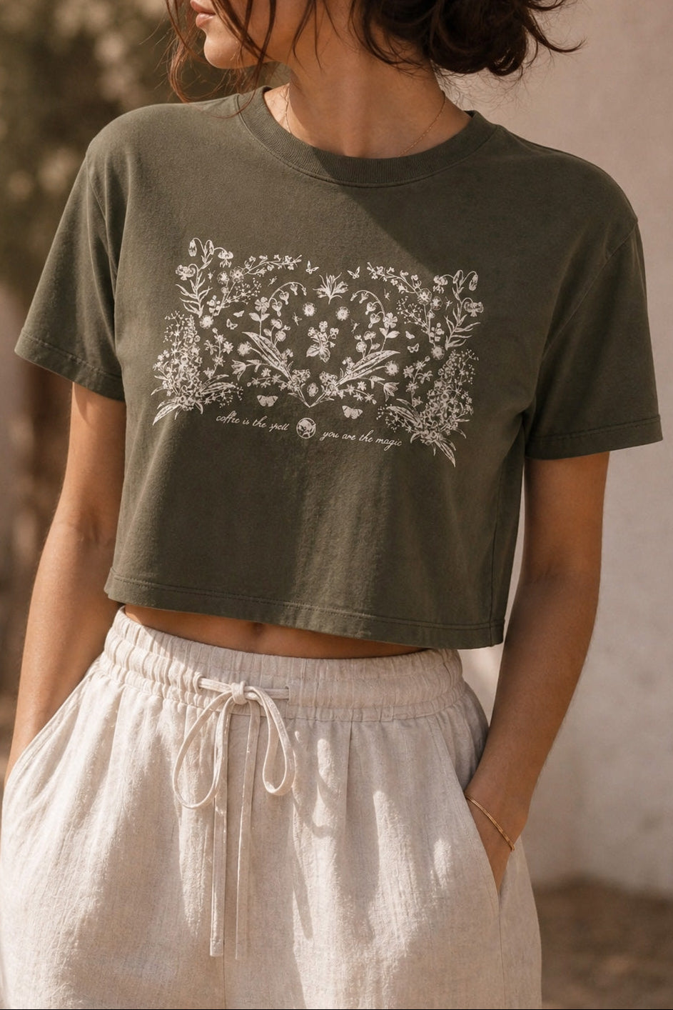 A woman wearing an olive green cropped t-shirt with a white floral graphic print on the front, paired with light beige drawstring pants, set against a warm, sunlit outdoor background.