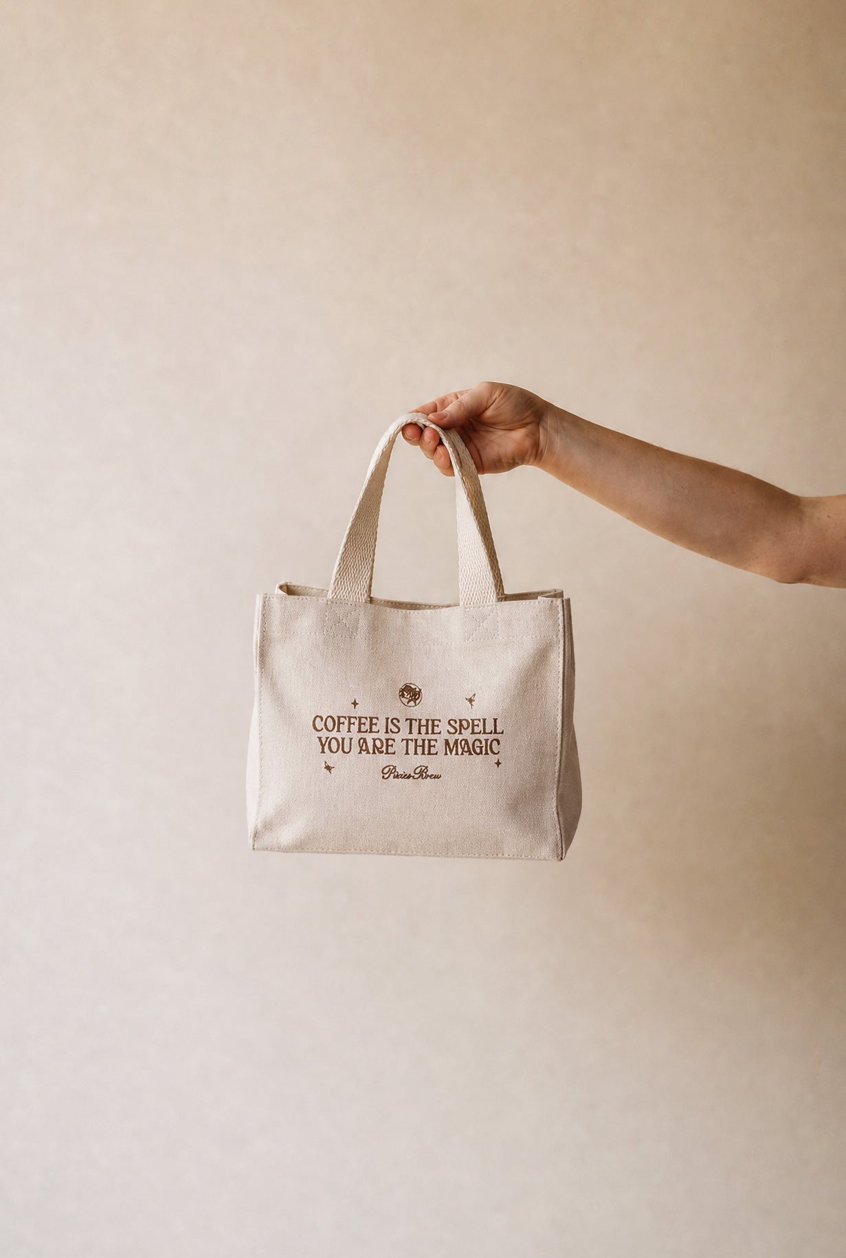A hand holding a small beige canvas tote bag with the text 'COFFEE IS THE SPELL YOU ARE THE MAGIC' and 'Chameleon' printed on the front, set against a plain beige background.