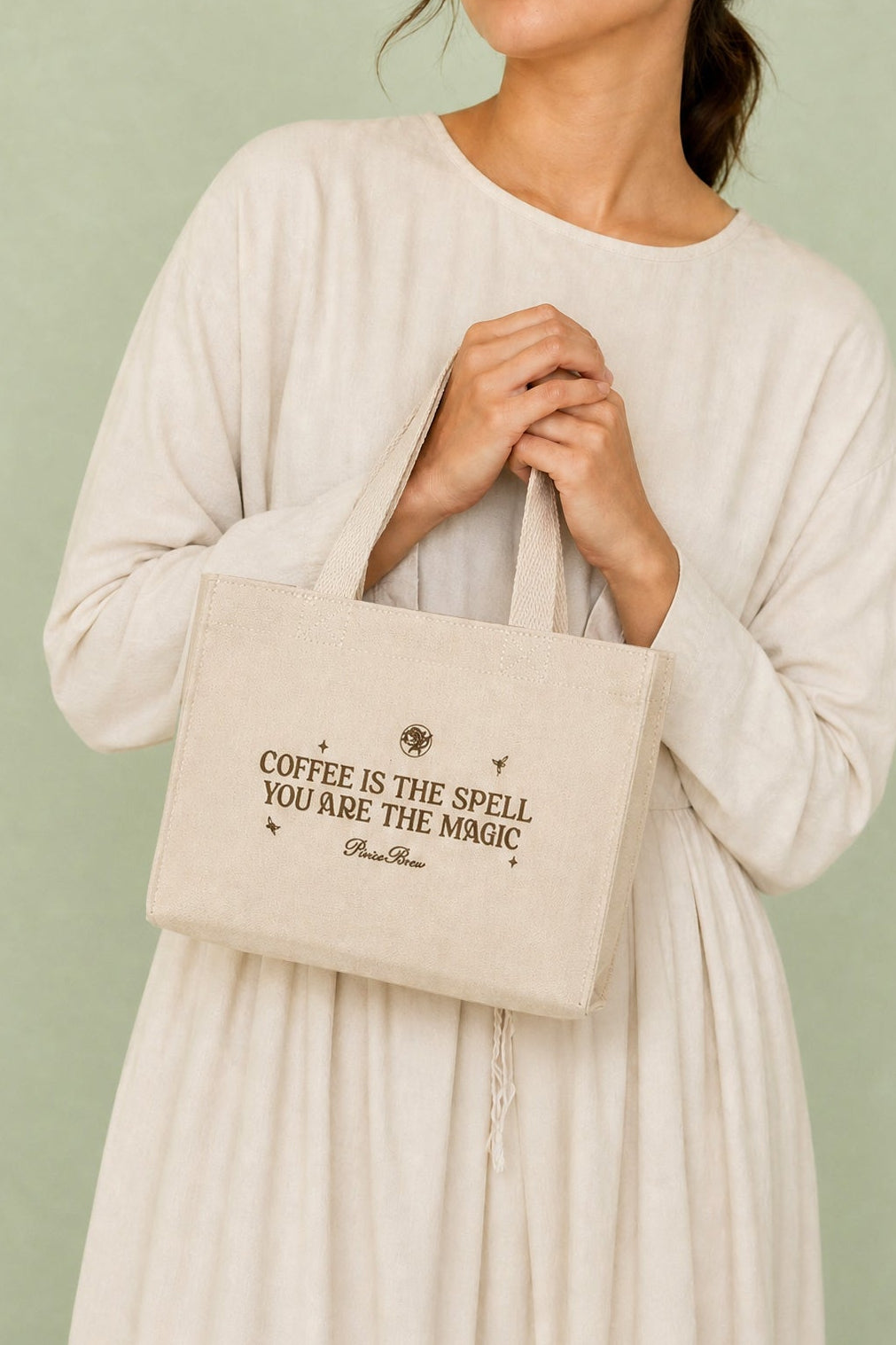A woman wearing a cream-colored long-sleeve dress holds a beige canvas tote bag with black text that reads 'COFFEE IS THE SPELL YOU ARE THE MAGIC'. The background is a solid sage green.