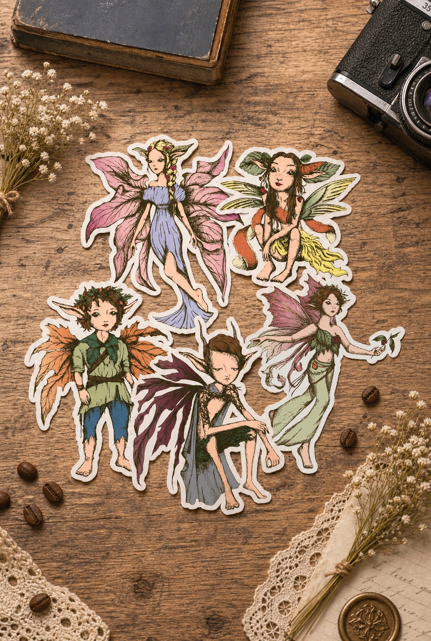 Five illustrated fairy and child character stickers arranged on a rustic wooden surface with dried flowers, lace, coffee beans, and a vintage camera nearby.