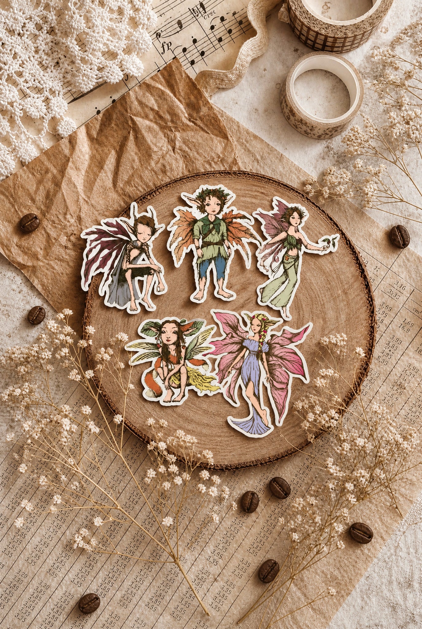 Five die-cut stickers featuring whimsical fairy characters arranged on a beige fabric background with dried flowers and soft shadows.
