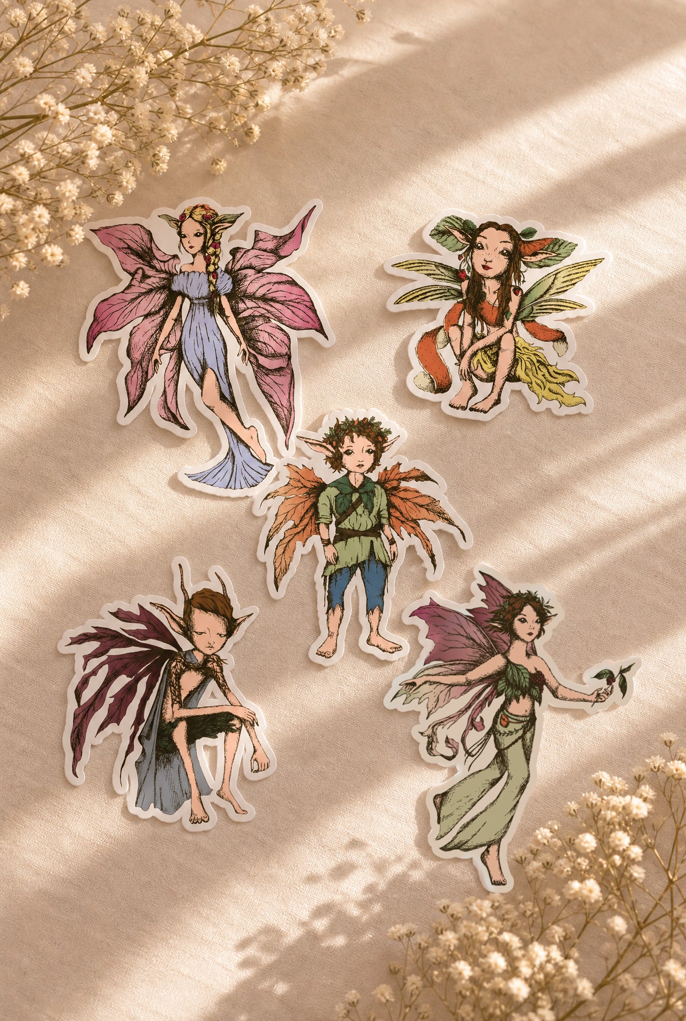 Five die-cut stickers featuring whimsical fairy characters arranged on a beige fabric background with dried flowers and soft shadows.